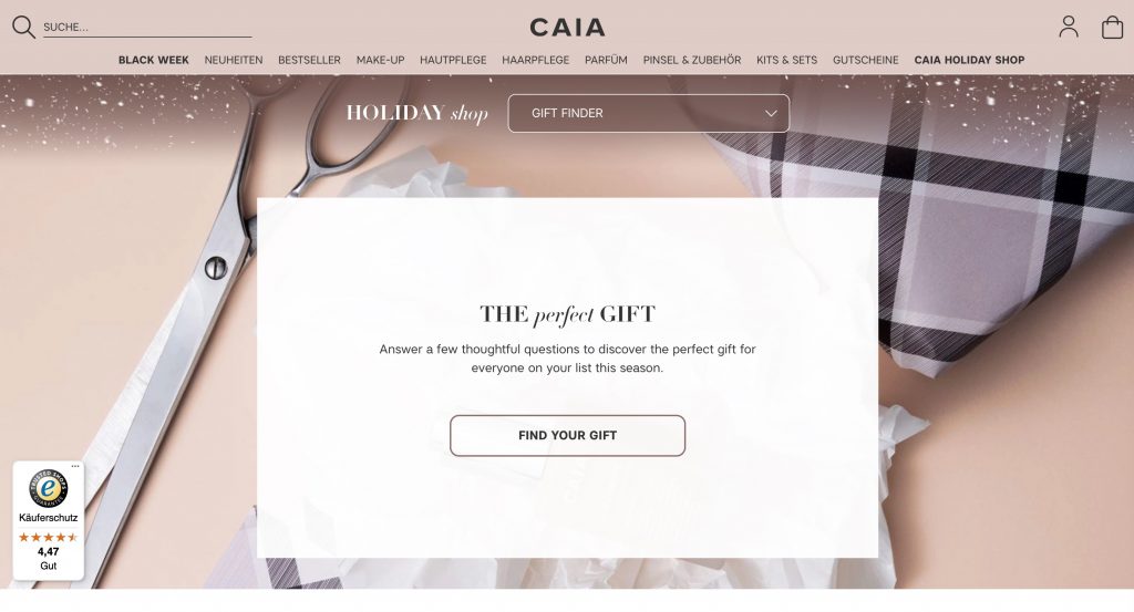 Screenshot der Homepage des Kosmetik- und Beauty-Shops CAIA. Die Navigation zeigt Kategorien wie "Black Week", "Make-Up", "Hautpflege", "Haarpflege", "Parfüm" und "Gutscheine". Im Zentrum der Seite ist ein großer Banner mit dem Titel "HOLIDAY shop" und dem Untertitel "THE perfect GIFT" zu sehen. Der Text fordert dazu auf, einige Fragen zu beantworten, um das perfekte Geschenk für die Saison zu finden. Darunter befindet sich ein Button mit der Aufschrift "FIND YOUR GIFT". Im Hintergrund sind eine große Schere und ein karierter Stoff sichtbar. Unten links wird das Trusted Shops Siegel mit der Bewertung "4,47 Gut" angezeigt.